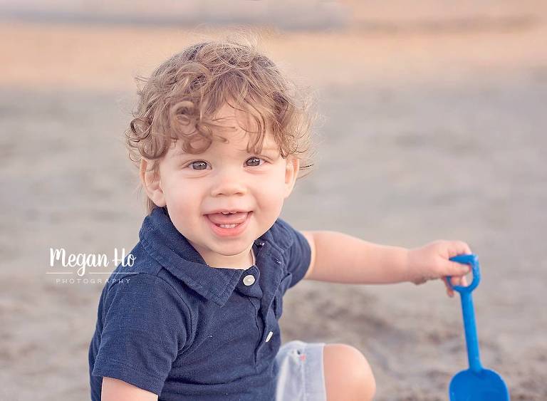 one year boy with brown curls smiling on nh beach with blue shovel