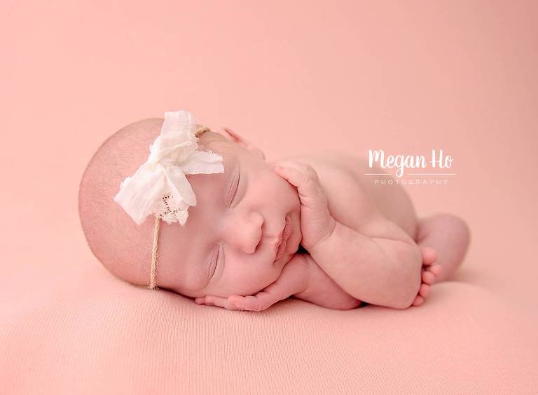beautiful baby girl with white bow on pink blanket cradling head in hands sleeping