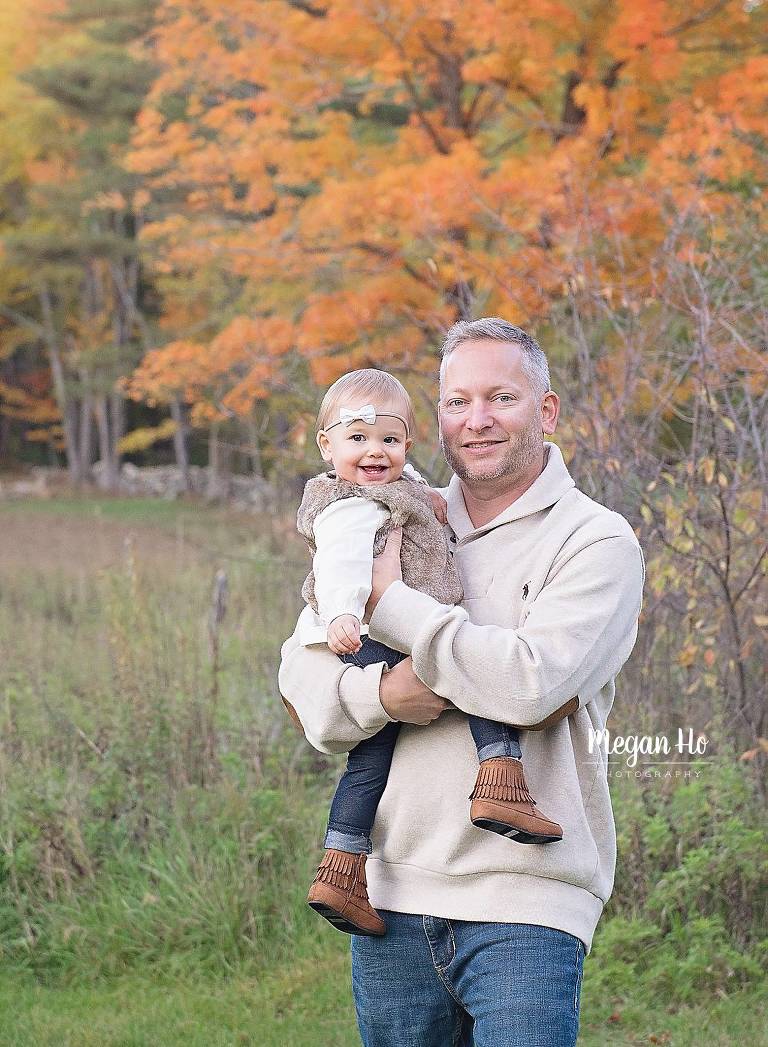dad holding one year old girl in nh fall session in front of orange leaves tree