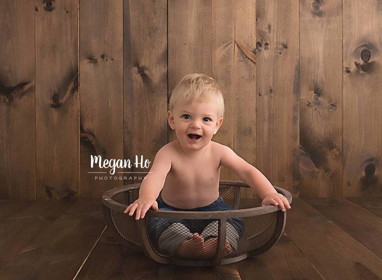 happy one year old boy in jeans sitting in wooden bowl