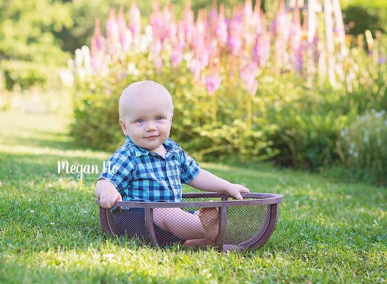 one year boy sitting in a mesh bowl in front of purple flowers