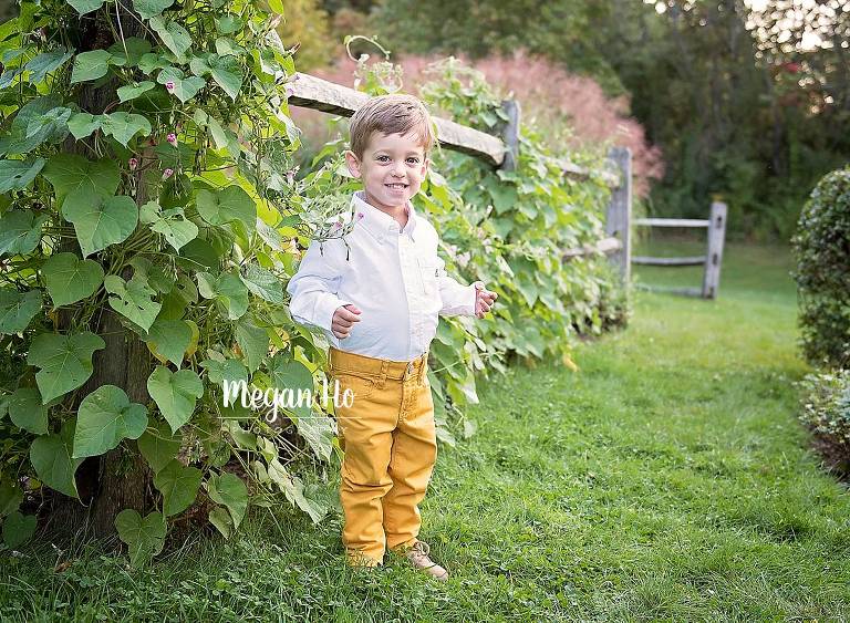 little two year old boy standing in front of ivy fence with yellow pants