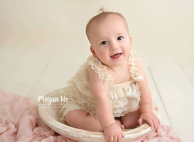 six month girl sitting in white bowl in cream lace romper with curl on top of head