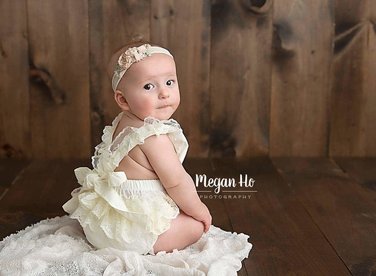 beautiful six month girl sitting on wood in bow romper with back rolls