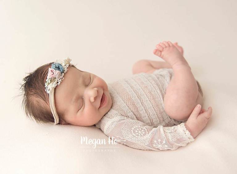 smiling baby girl in lace romper with bow headband on cream blanket