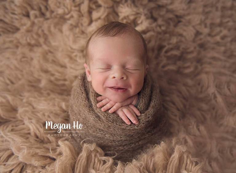 baby boy smiling wrapped in brown wrap on tan flokati rug in newborn session