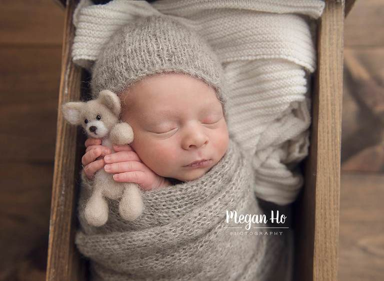 sleeping newborn snuggling little bear wrapped in grey with bonnet on in little wood bed