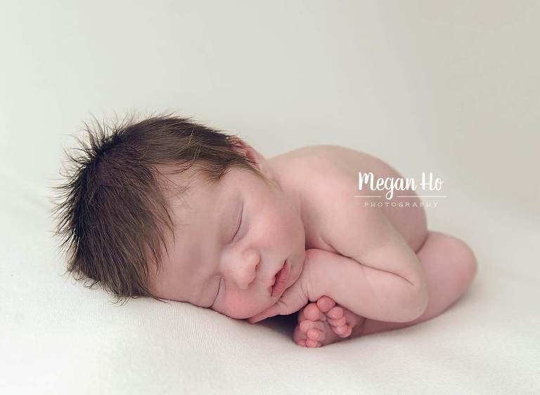 curled up newborn boy sleeping with hands on toes on white backdrop