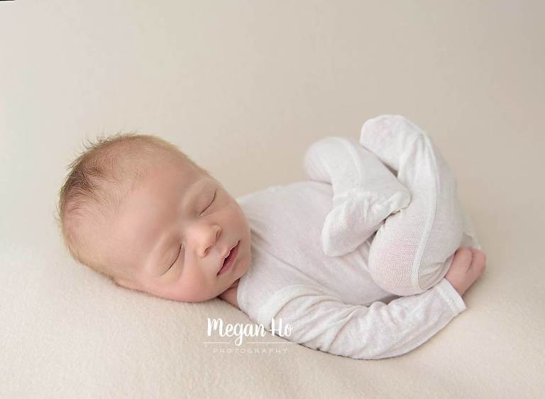 baby boy on tan in white outfit with feet folded up sleeping on back