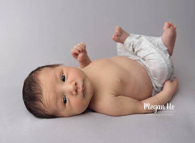 awake newborn baby with brown hair in grey pants looking at camera