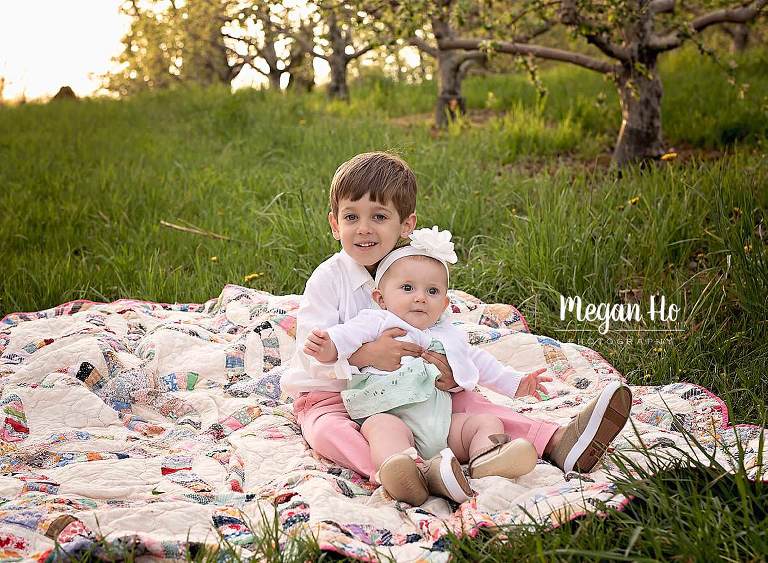Big brother happily holding little sister on quilt dressed in white, pink and green