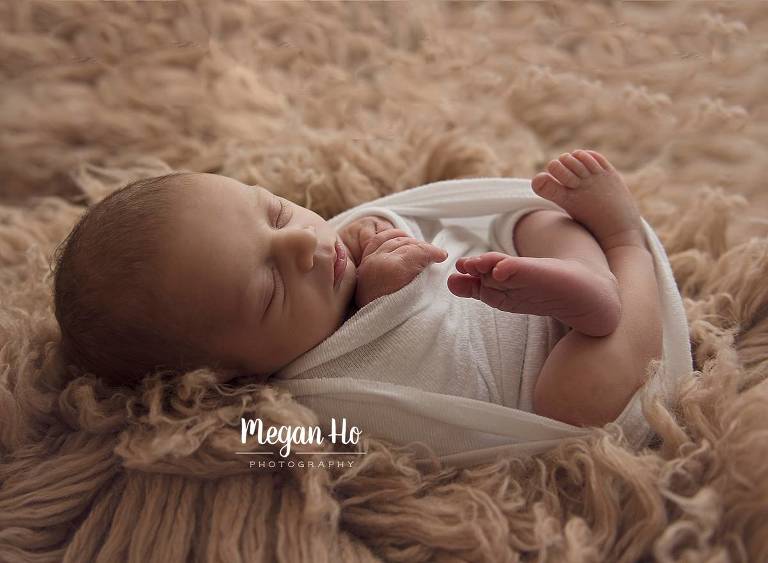 infant boy wrapped in white on tan rug sleeping backlit with light