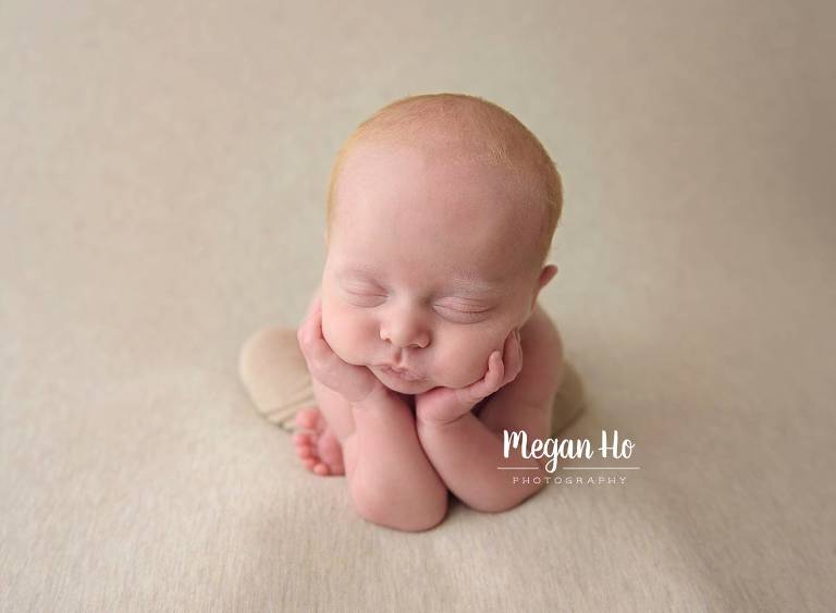 little red head baby boy head in hands on tan backdrop in little pants