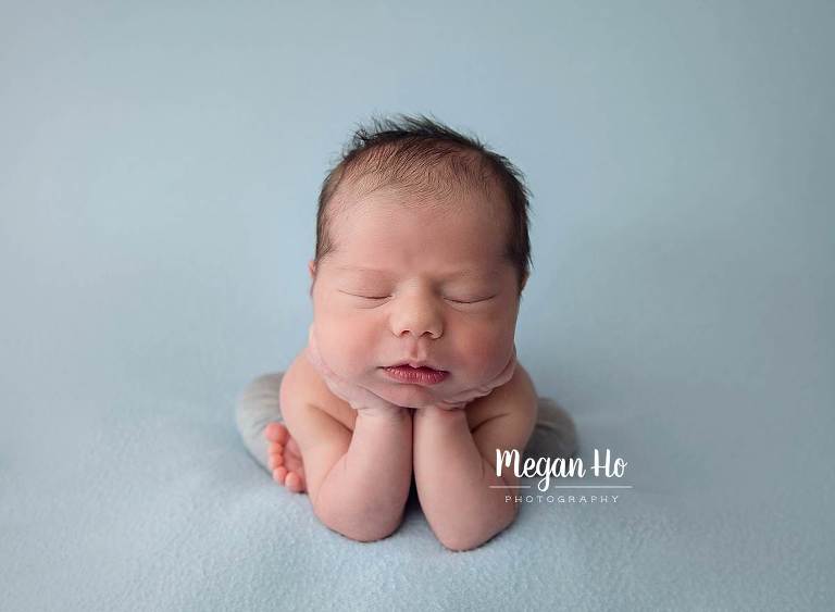 newborn baby boy froggy pose with head in hands in little grey pants on blue