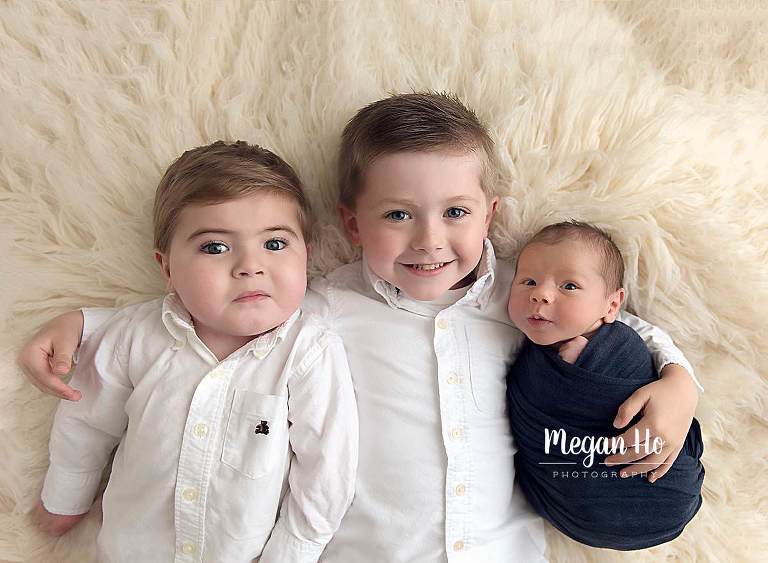 three smiling brothers laying snuggled together on white fluffy rug in newborn session