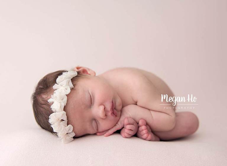 adorable curled up baby girl with white bow headband on pink blanket